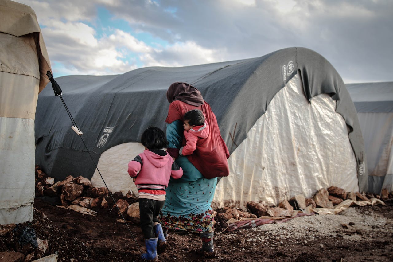 who-we-are A woman and children walk near tents in a refugee camp in Idlib, showcasing resilience amidst adversity.