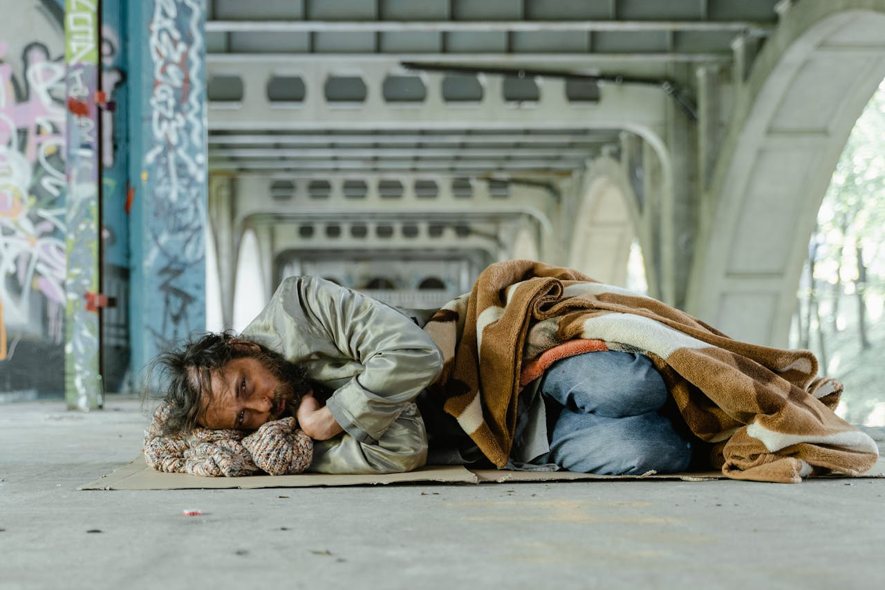 services-03 A homeless man lies under an urban bridge, wrapped in a blanket, symbolizing poverty and survival.